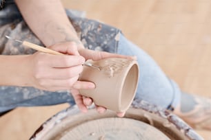 Mãos de jarro feminino jovem ou ceramista com padrões de escultura de ferramentas manuais nas laterais do copo de barro artesanal enquanto sentado pela roda de cerâmica