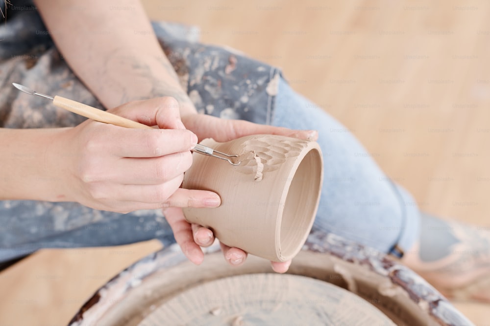 陶器のろくろのそばに座っている手作りの粘土カップの側面に手工具の彫刻パターンを持つ若い女性のピッチャーまたは陶芸家の手