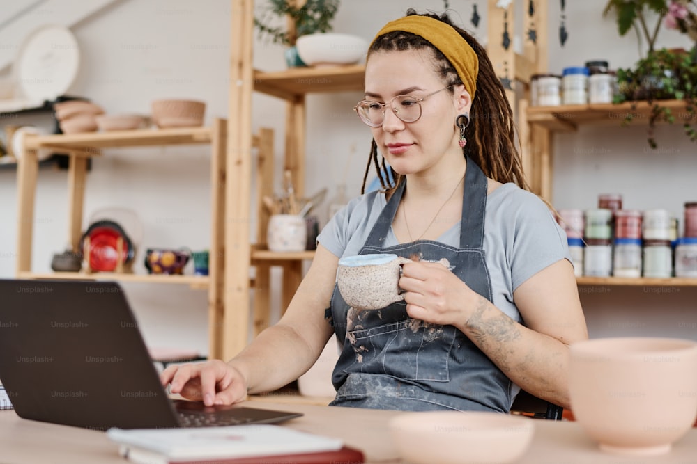 Young female freelancer or owner of small business sitting in front of laptop by workplace, having tea and searching for new ideas in the net