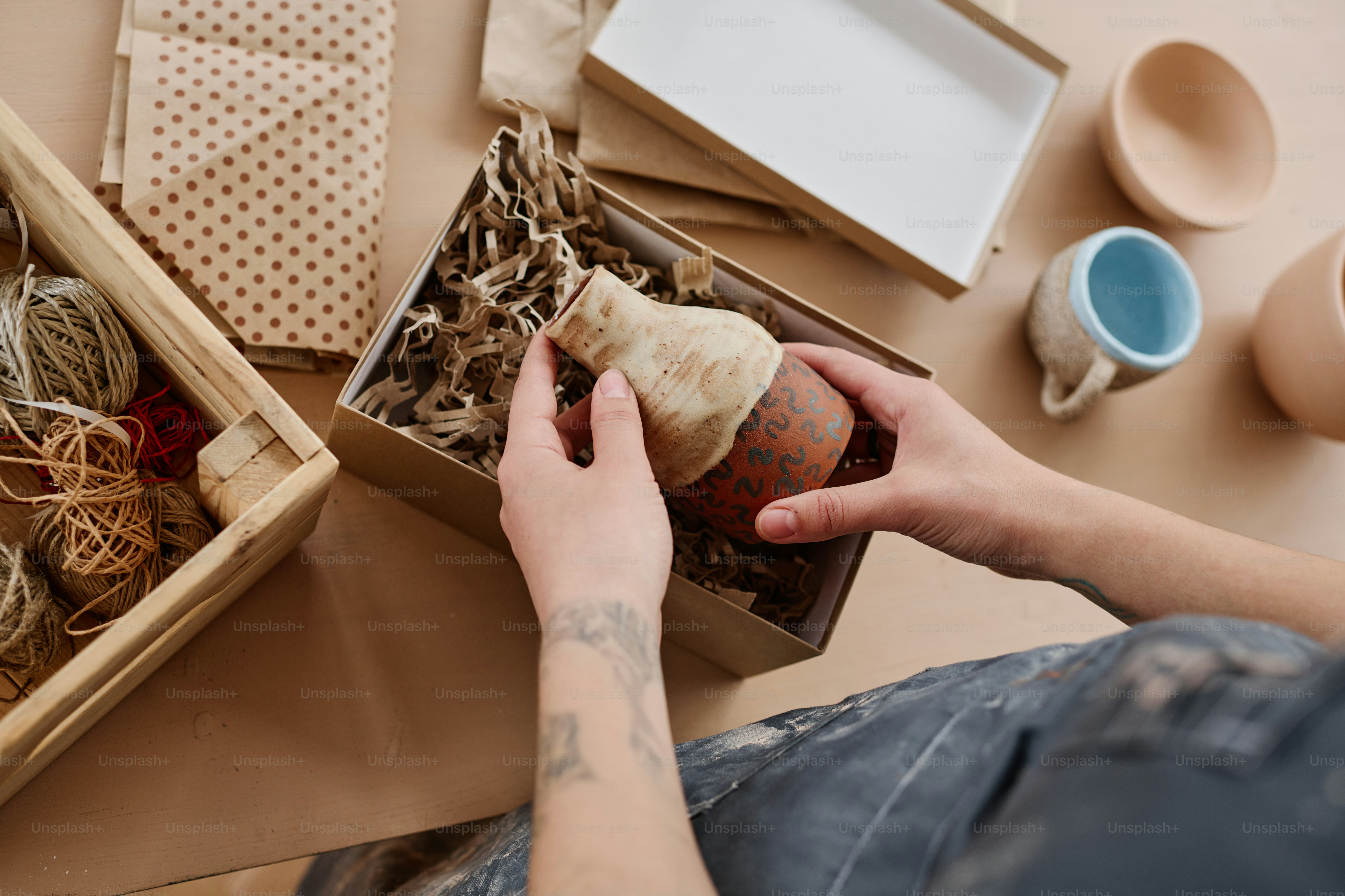 Hands of young female pottery master putting handmade jug into cardboard box with bast fiber or cut paper by workplace