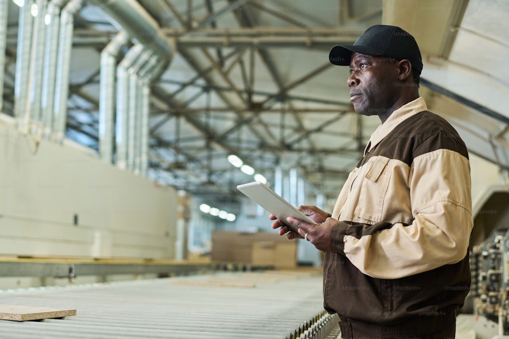 Ernsthafter afrikanischer Bauarbeiter in Uniform, der an einem digitalen Tablet arbeitet, während er in der Werkstatt der Fabrik steht