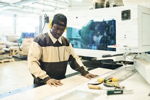 African carpenter in uniform standing at machine and cutting wooden board during production of furniture