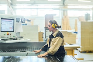 Jovem trabalhadora em fones de ouvido de proteção em pé na máquina na fábrica