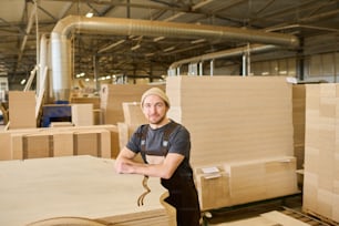 Portrait of young worker in overalls looking at camera standing at warehouse near the wooden products