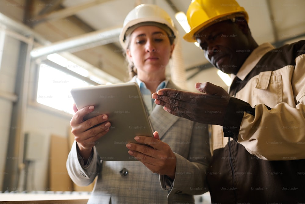 Caposquadra africano con elmetto che indica il tablet pc e parla con l'ingegnere durante il lavoro in fabbrica