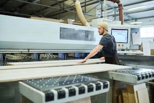 Young bearded worker standing at modern computer machine and working with wooden planks at factory