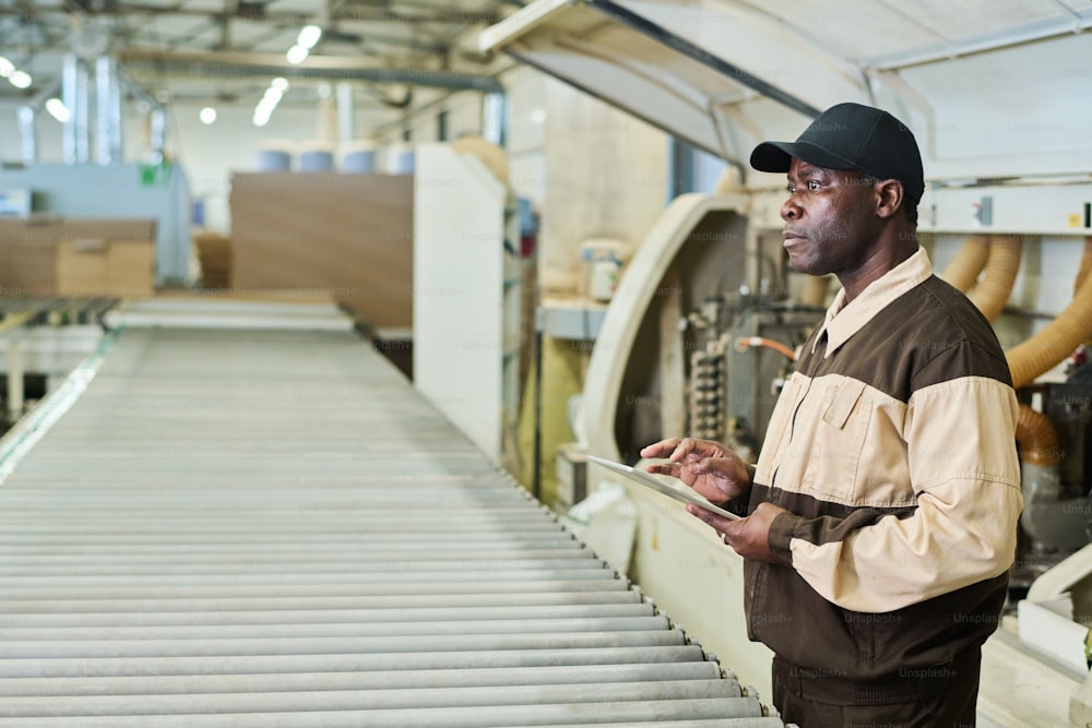 Afrikanischer Vorarbeiter in einheitlicher Steuerungsmaschine mit digitalem Tablet während seiner Arbeit in der Fabrik