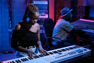 スタイリッシュな若いアフリカ系アメリカ人女性ミュージシャンがデジタルピアノを弾き、オーディオエンジニアがレコーディングスタジオでミキサーで作業する
