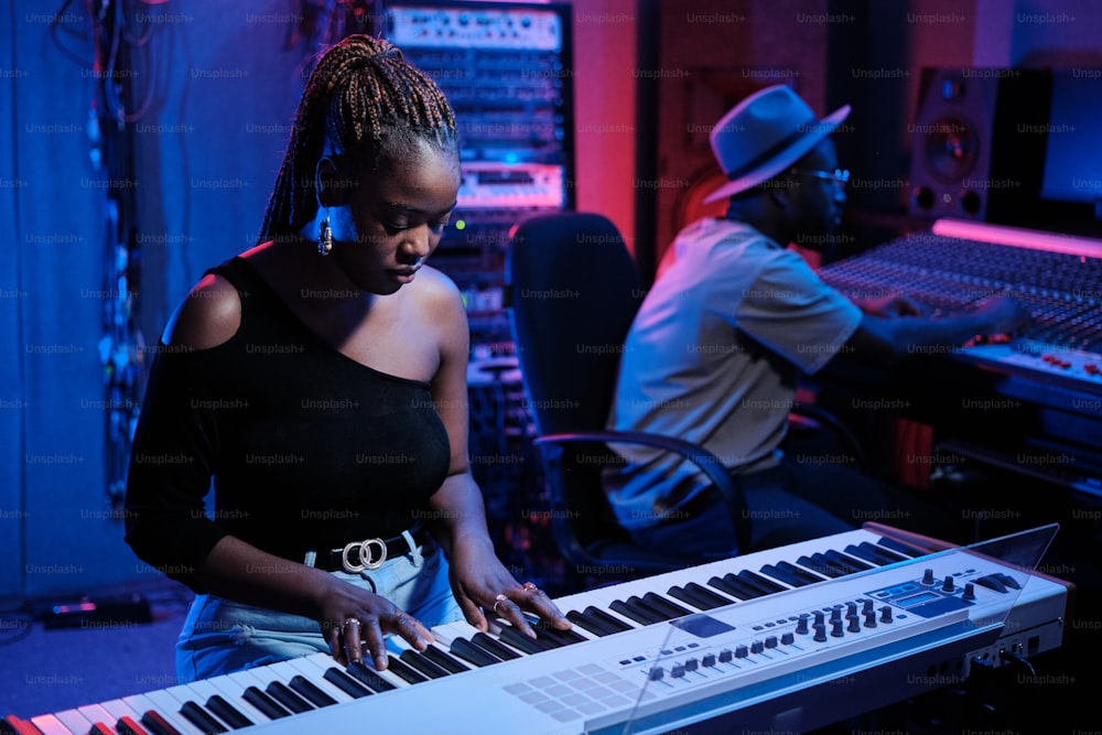 Stylish young African American female musician playing digital piano while audio engineer working on mixer in recording studio