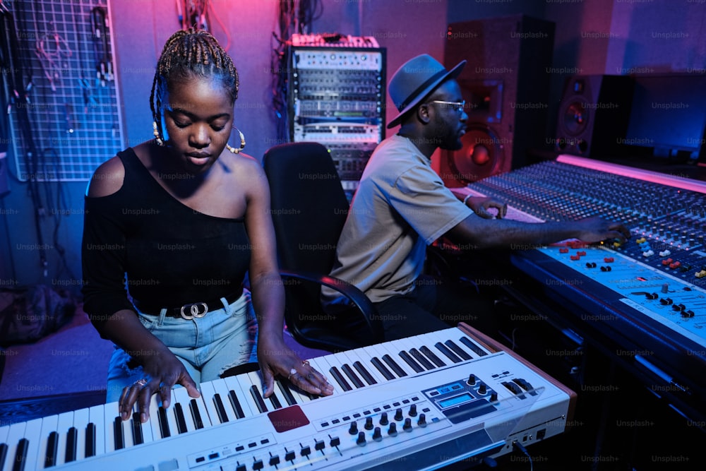 Modern young African American female musician playing digital keyboard while sound engineer working on mixer in recording studio