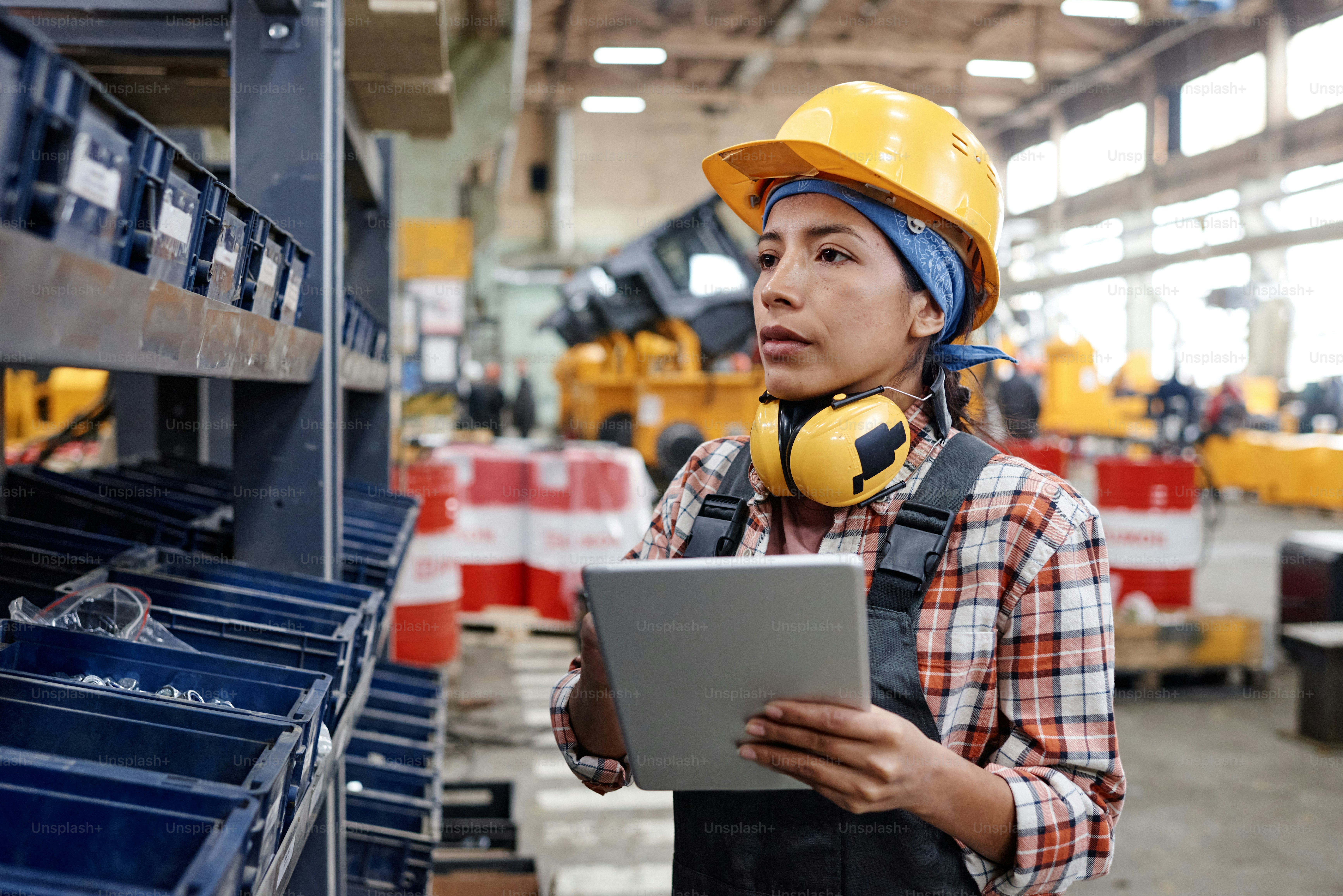 Young female with tablet standing by shelf with large plastic containers with bolts and other steel stuff necessary for machinery production