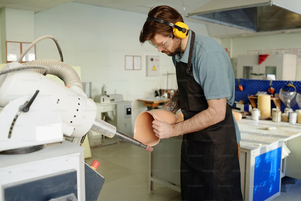 Junge Manufakturarbeiterin in schützenden Kopfhörern und Brillen glättet Kanten der Silikon-Sockelauskleidung in der Werkstatt