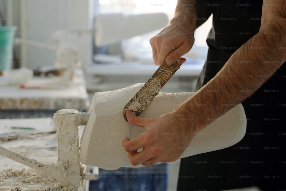 Mains d’un jeune ouvrier d’usine avec un outil à main enlevant le plâtre excessif de la surface du plâtre tout en se tenant près de l’établi
