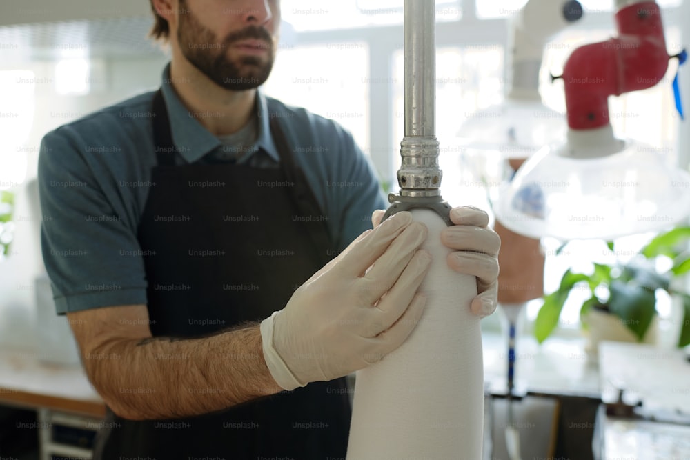 Handschuhe junger männlicher Spezialist, der thermoplastische Platten über dem Prothesenschaft mit Hilfe eines speziellen Metallkolbens in der Werkstatt befestigt
