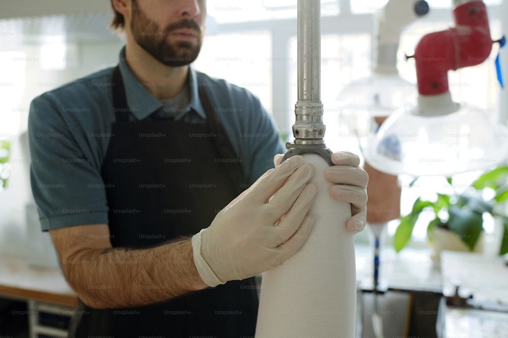 Gloved young male specialist fixing thermoplastic sheet over prosthetic socket with help of special metallic piston in workshop