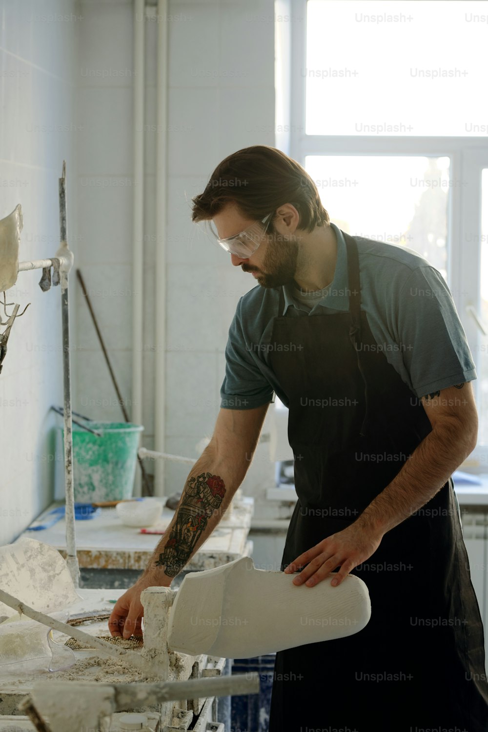 エプロンを着た若いひげを生やした労働者が作業台のそばに立ち、クライアントのために新しい石膏模型を処理するための手工具を取る