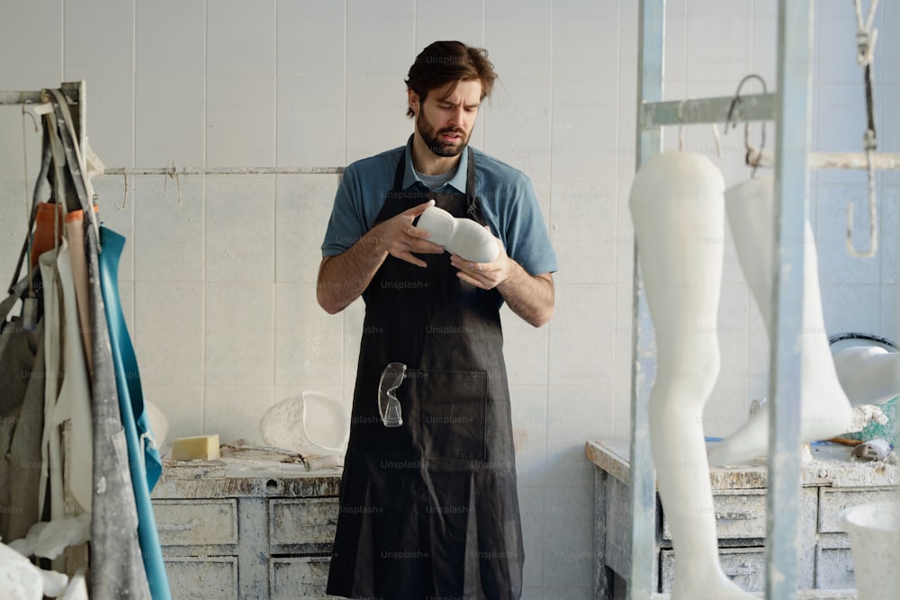 Junger Meister der prothetischen Manufaktur betrachtet Gipsabdruck in seinen Händen, während er vor einem ausgestatteten Arbeitsplatz steht