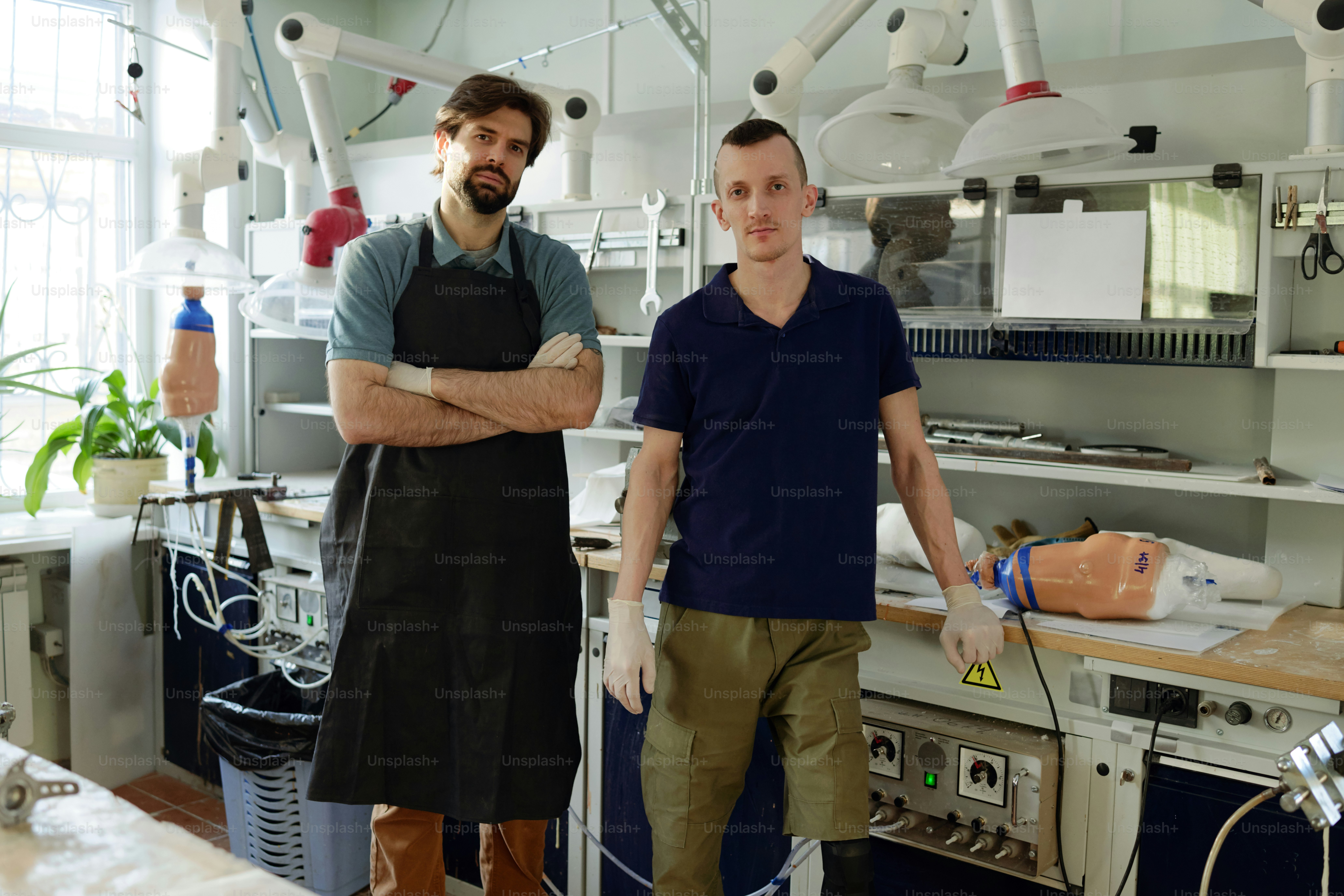 Dois jovens especialistas em vestuário de trabalho em pé na grande oficina de fabricação de próteses e olhando para a câmera