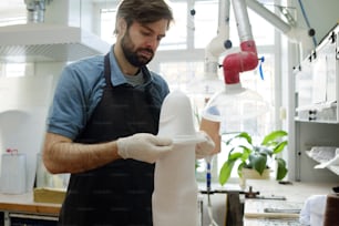 Young serious man in apron and gloves putting thermoplastic sheet all over prosthetic socket while working over one of clients orders