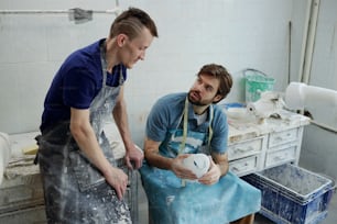 Two young workers of prosthetic factory in aprons discussing working points while one of them holding socket for prosthesis