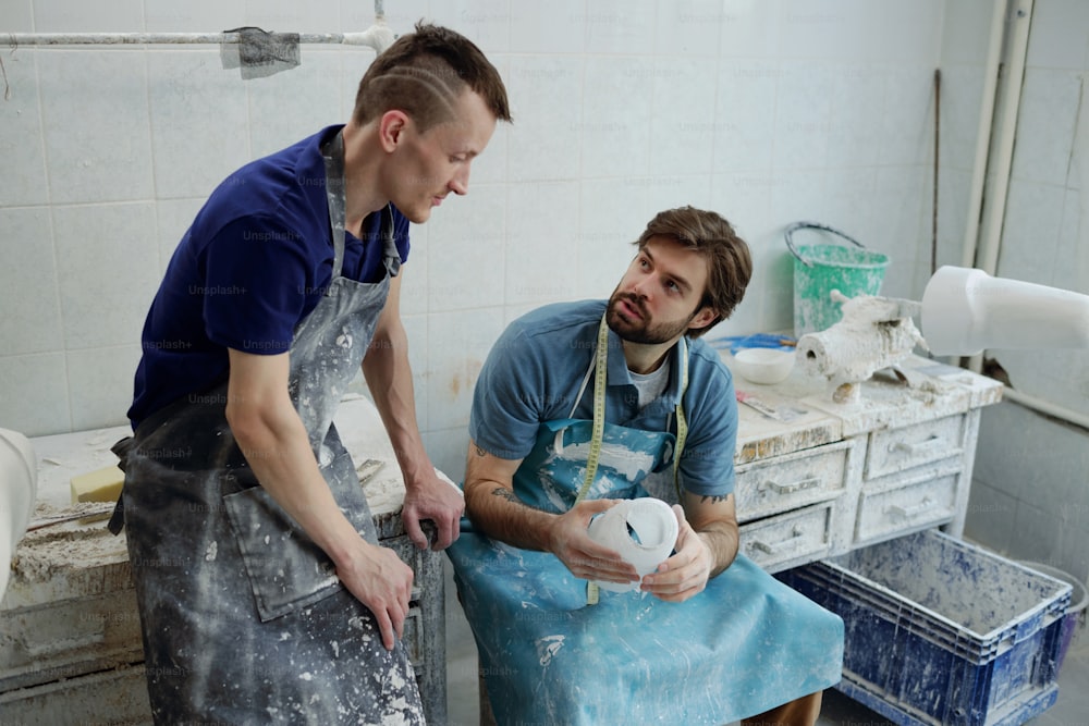 Two young workers of prosthetic factory in aprons discussing working points while one of them holding socket for prosthesis