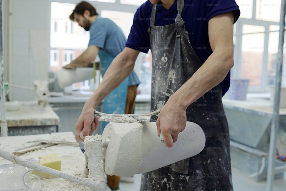 Young worker in apron holding worktool while grinding plaster cast of part of prosthetic limb on background of coworker