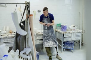 Young man in apron making revision of work supplies in prosthetic factory workshop while standing by table with plaster casts