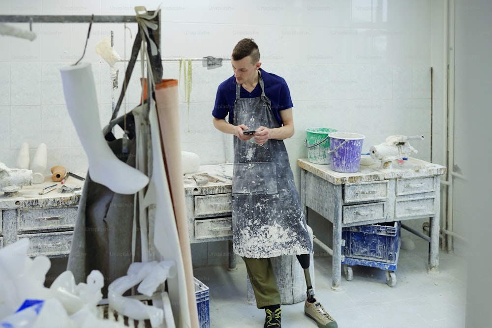 Giovane uomo in grembiule che fa revisione delle forniture di lavoro nell'officina della fabbrica di protesi mentre sta in piedi dal tavolo con i calchi in gesso