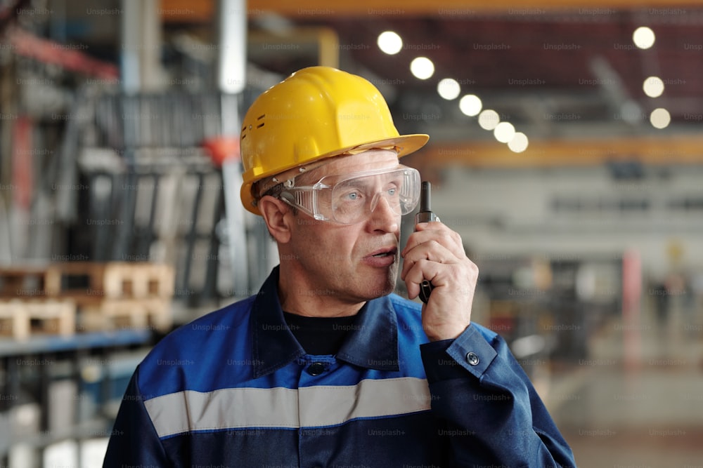 Confident mature Caucasian factory foreman in hardhat and safety goggles using radio device to communicate with coworkers