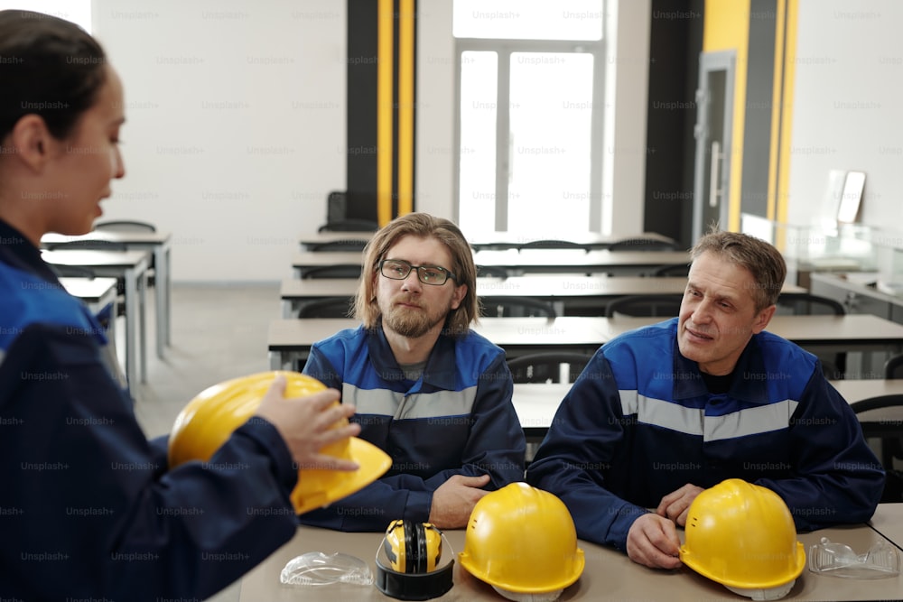 Arbeiter, die mit Schutzhelmen und Schutzbrillen in der Fabrikkantine am Tisch sitzen und miteinander plaudern