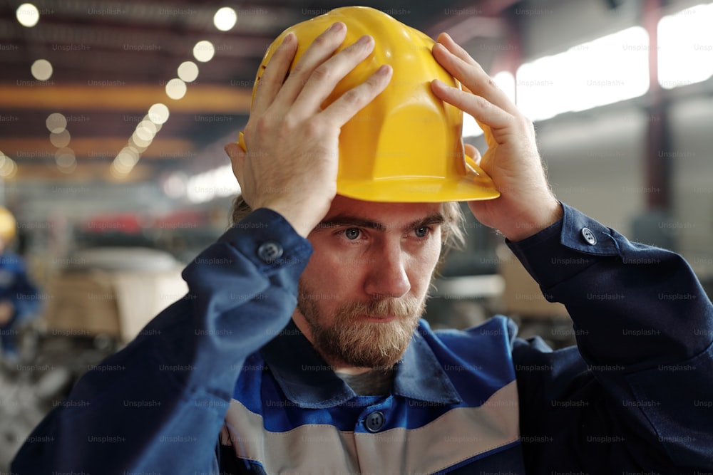 Serious young bearded man standing at factory and putting hardhat on head while preparing for work
