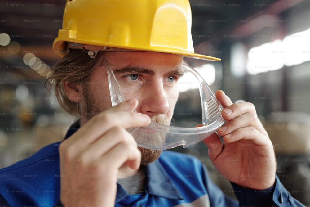 Close-up of confident young bearded factory worker in yellow hardhat putting safety goggles on face while preparing for work
