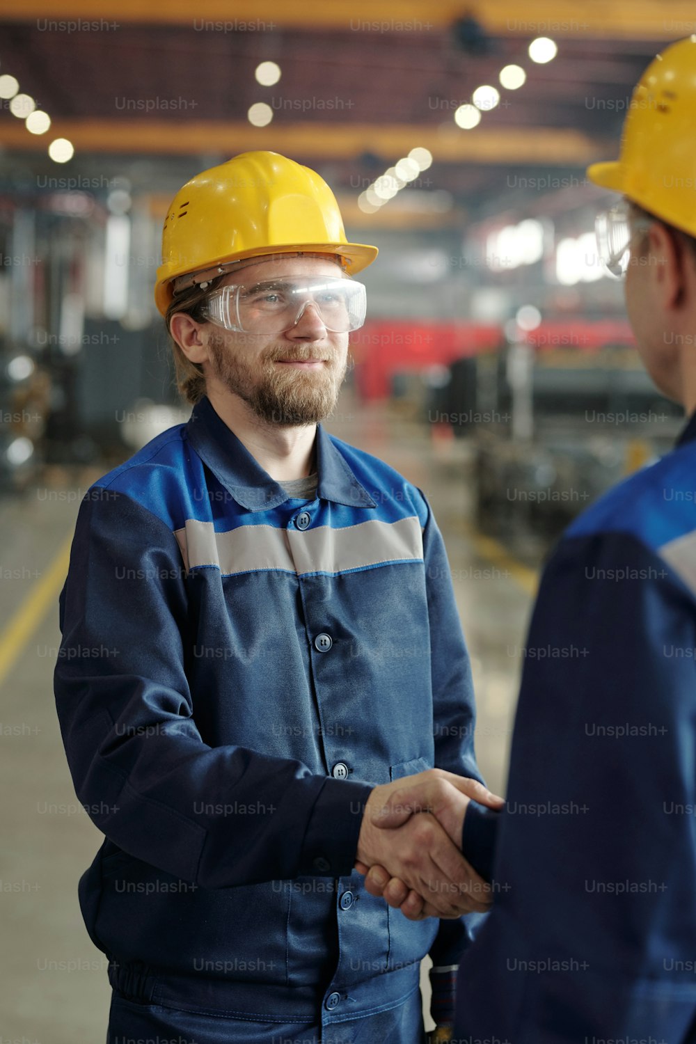 Smiling handsome bearded Caucasian employee in hardhat and goggles shaking hand of coworker at factory