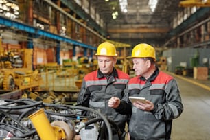 Two workers in helmets using tablet pc and discussing the work of machine equipment in the plant