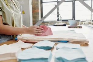 Hände einer jungen Schneiderin, die rosa Stoffwerkstücke für Schulterpolster über einem Stapel gefalteter Textilien auf einem großen Tisch in der Werkstatt hält