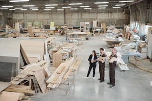 Young worker of furniture factory in uniform showing female manager in formalwear new equipment at working meeting in warehouse