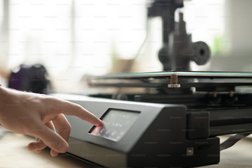 Mão do jovem designer tocando botão no painel de controle da impressora 3D enquanto vai imprimir novos itens no escritório
