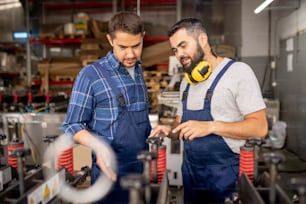 One of two factory workers explaining his colleague how to use new industrial equipment and pointing at detail