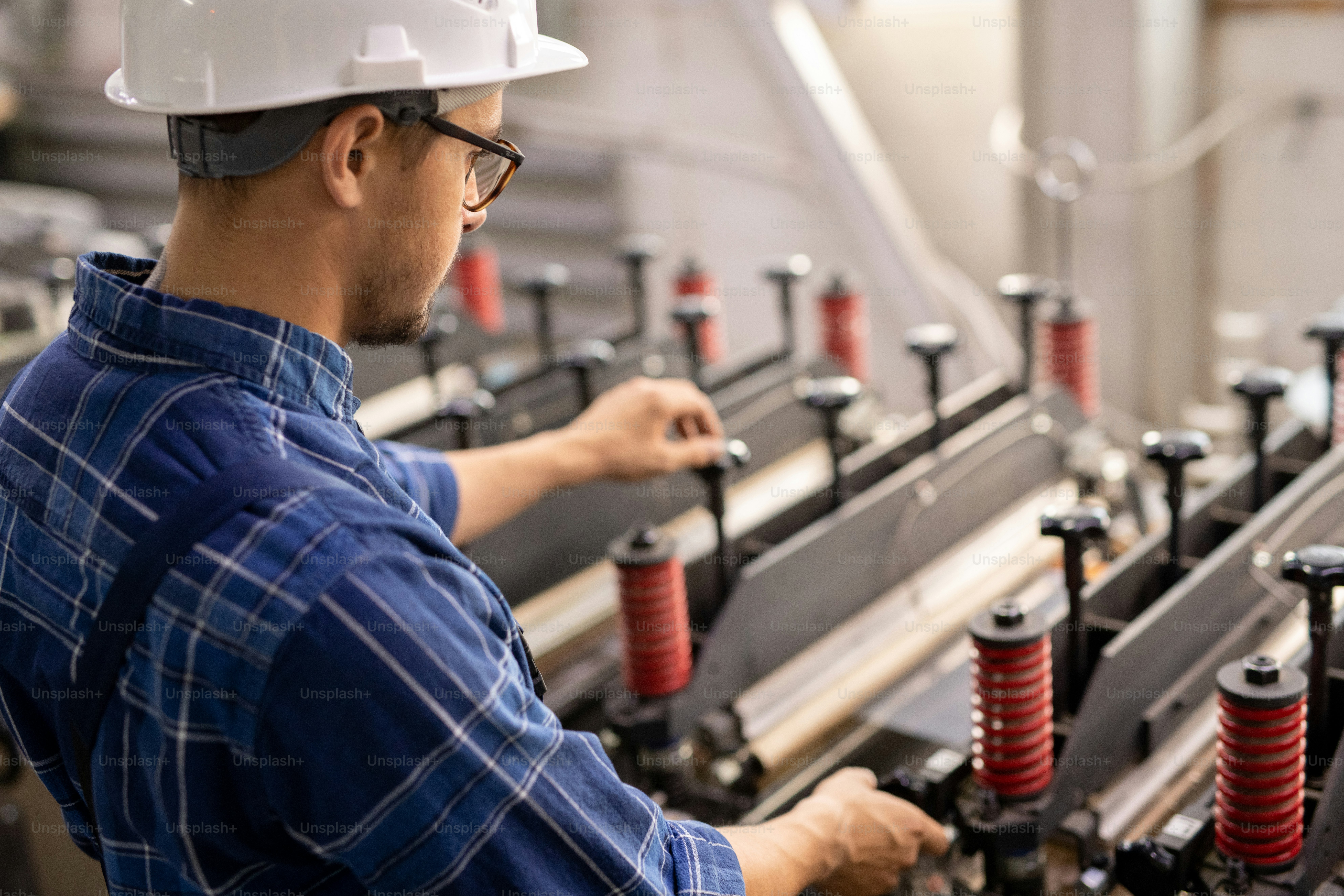 Young engineer in white helmet standing by industrial machine while operating it in working area