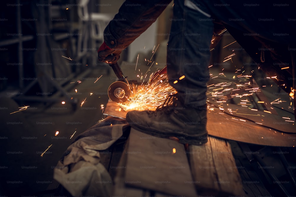Vista lateral de perto do trabalhador de tecido trabalhando com a ferramenta de moagem elétrica em uma estrutura de aço na fábrica enquanto faíscas voam.