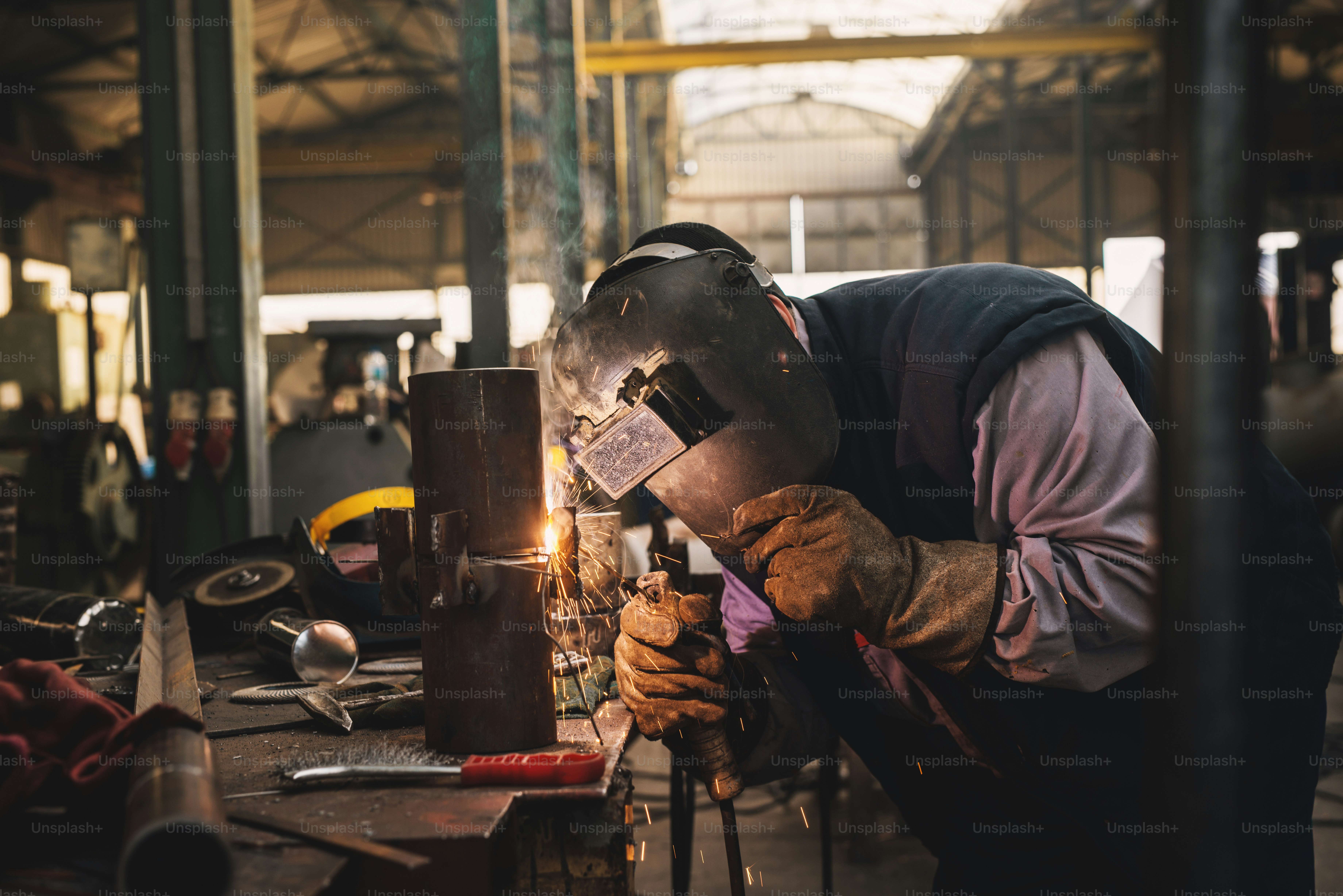 Soldar em uniforme de proteção e máscara de solda tubo de metal na mesa industrial enquanto faíscas voam.