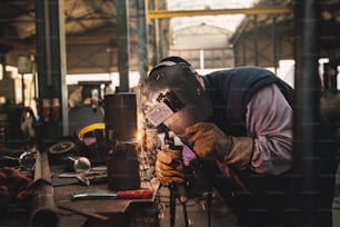 Welder in protective uniform and mask welding metal pipe on the industrial table while sparks flying.