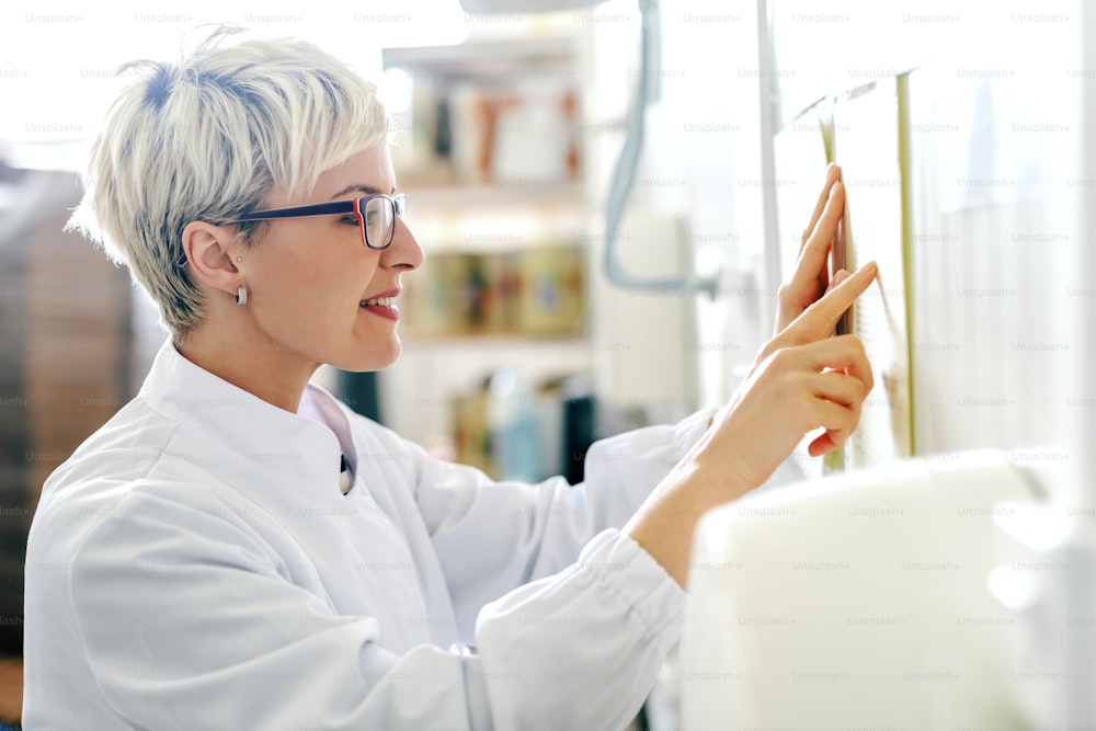 Profile of young Caucasian blonde female employee reading schedule on the wall while standing in food factory.