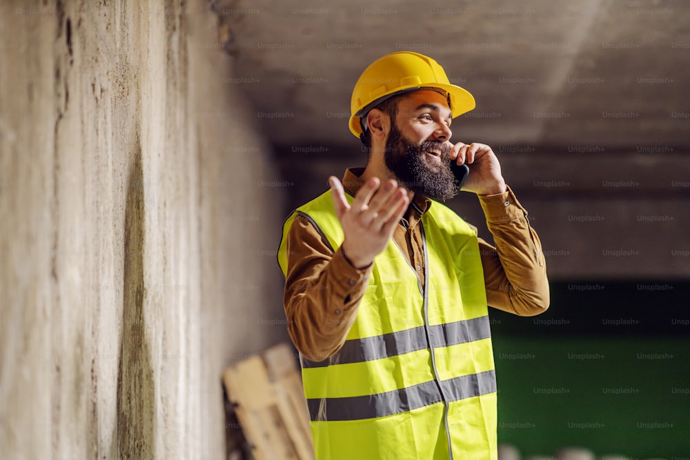 Jovem trabalhador barbudo bonito em pé na frente da parede de concreto e falando ao telefone inteligente.