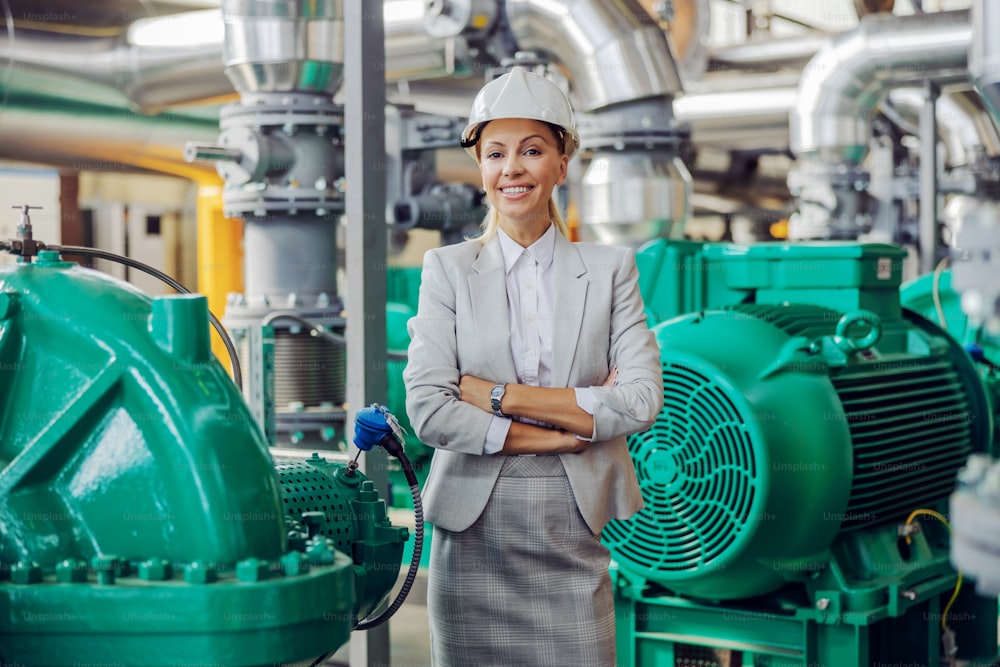 Middle aged successful independent female CEO in suit with helmet on head standing in heating plant with arms crossed and looking at camera.