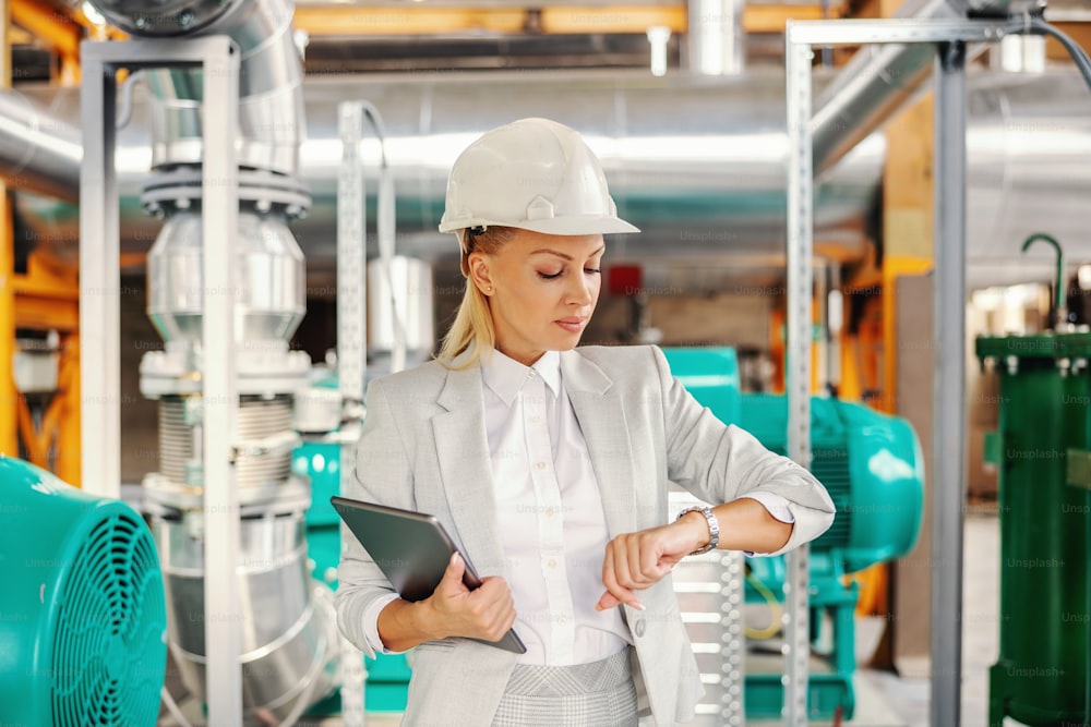 Middle aged successful blond female supervisor in suit with helmet on head holding tablet and looking at wristwatch and waiting for a meeting while standing in heating plant.