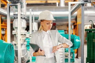 Middle aged successful blond female supervisor in suit with helmet on head holding tablet and looking at wristwatch and waiting for a meeting while standing in heating plant.