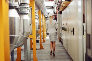 Rear view of experienced independent female supervisor in suit with face mask and helmet walking around heating plant and looking around during corona virus outbreak.