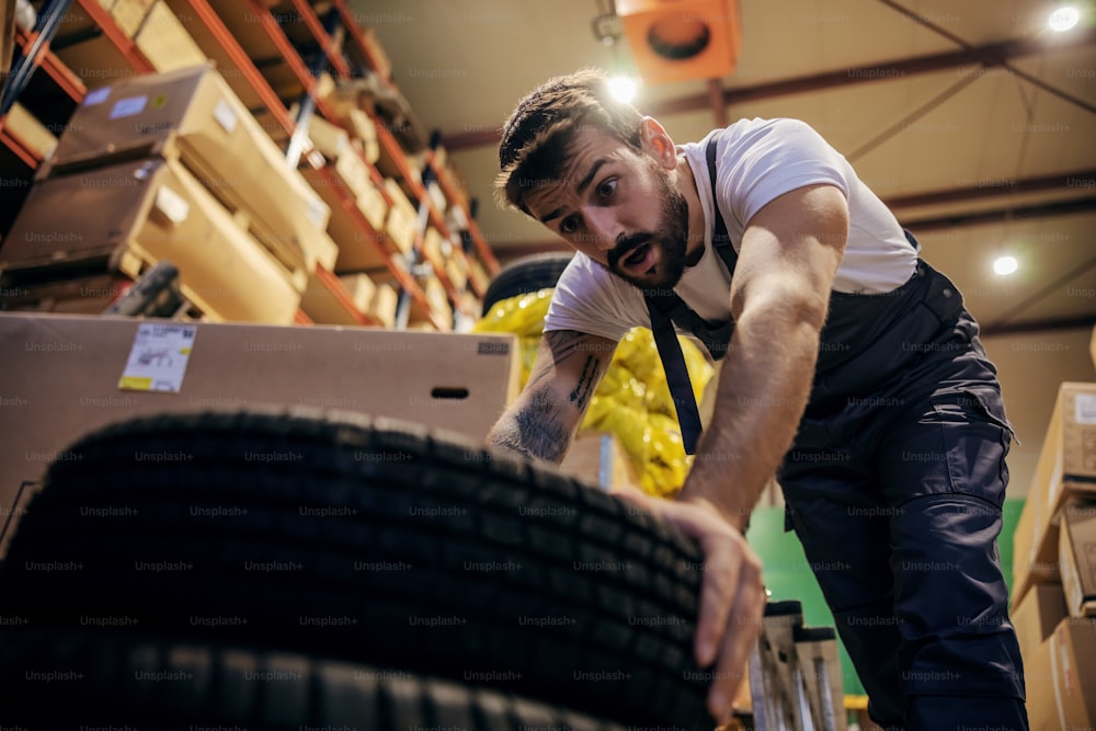 Bearded tattooed worker trying to lift tires in storage of import and export firm.