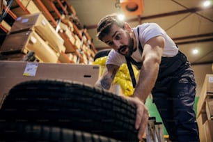 Bearded tattooed worker trying to lift tires in storage of import and export firm.